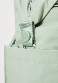 Mochila de tela verde suave con un cierre de botón texturizado y correas duraderas. Presenta un bolsillo lateral y acentos de costura suave.