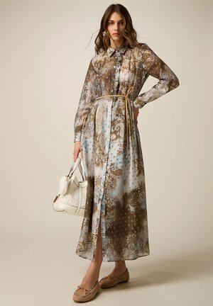 Kvinde iført en langærmet, mønstret knælængde kjole med tyndt bælte, holder en hvid håndtaske og har på brune mokkasiner.