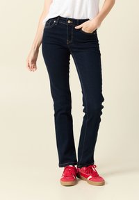 Ciemnoniebieskie jeansy o dopasowanym kroju, z złotymi przeszyciami, pięcioma kieszeniami, z przodu zapinane na guzik, noszone z czerwonymi trampkami.