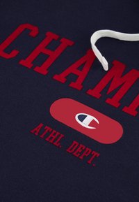 Felpa blu navy con scritte rosse in grassetto "CHAMP" e "ATHL. DEPT." Stringa bianca per il cappuccio. Presenta un logo dentro un ovale rosso.