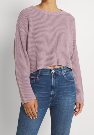Femme portant un pull court côtelé couleur lavande et un jean bleu taille haute, debout devant un fond clair uni.