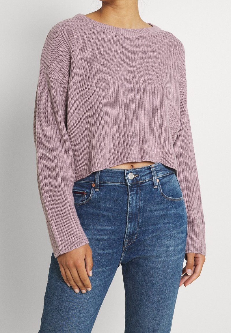 Femme portant un pull court côtelé couleur lavande et un jean bleu taille haute, debout devant un fond clair uni.