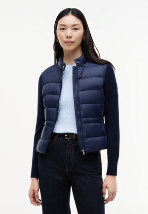 Mujer con chaleco acolchado azul marino sobre un jersey de punto de ochos azul claro y vaqueros oscuros, de pie frente a un fondo blanco liso.