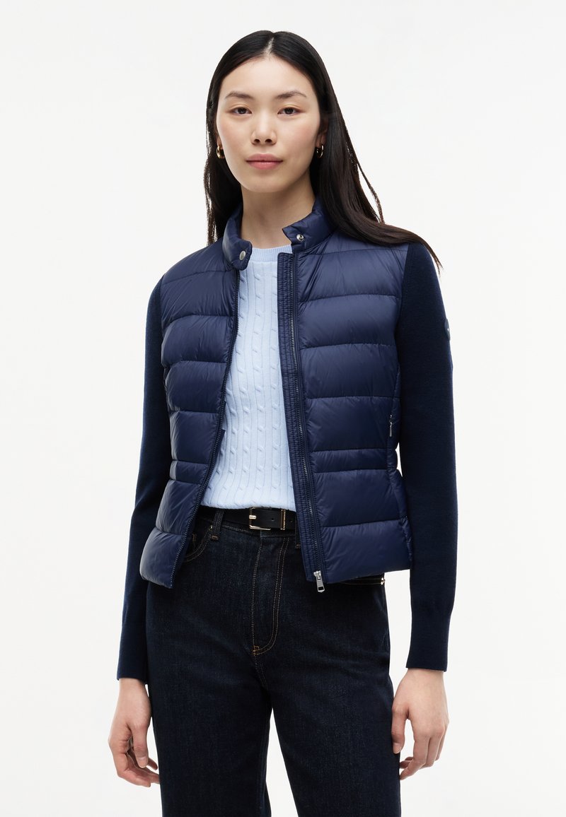 Femme portant un gilet matelassé bleu marine sur un pull à torsades bleu clair et un jean foncé, debout devant un fond blanc uni.