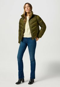 Oliwkowa pikowana kurtka z wysokim kołnierzem, zapinana na zamek, z ściągaczami przy rękawach, noszona na kremowym swetrze w splot warkoczowy i niebieskich dżinsach. Czarne botki na obcasie.