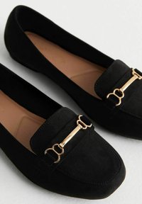 Mocassins en daim noir avec une semelle plate, présentant un accent en métal doré à l'avant et un design à bout arrondi. Texture lisse.