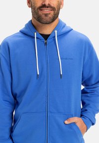 Blue zip-up hoodie made of cotton blend fabric. Features a hood, white drawstring accents, and front pockets. Embroidered logo on chest.