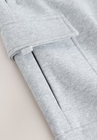 Gros plan d'une manche en tissu gris clair avec un poignet et une poche latérale inclinée avec un petit détail de boucle en tissu.