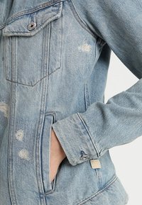 Jasnoniebieska kurtka dżinsowa o lekko zniszczonej fakturze, z przednimi kieszeniami, srebrnymi zapięciami na guziki i małą metką marki po bokach.