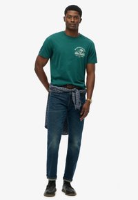 Zielona bawełniana koszulka z nadrukiem, zestawiona z ciemnymi dżinsami. Na talii przewiązana jest koszula w kratę. Całość dopełniają czarne buty.