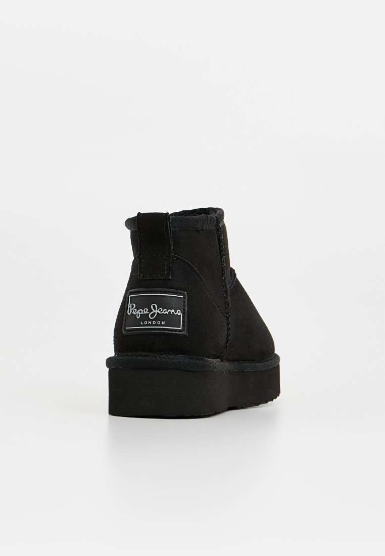 Schwarze Wildleder-Stiefeletten mit flacher Sohle, abgerundeter Spitze und einem auffälligen Logopatch an der Ferse, mit gestickten Akzenten und einer strukturierten Oberfläche.