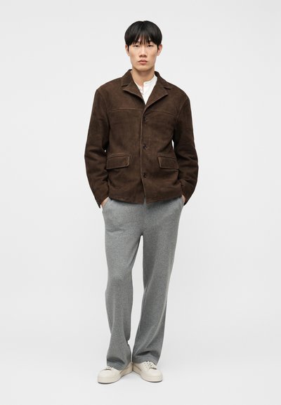 Άνδρας στέκεται με τα χέρια στις τσέπες, φορώντας καφέ σουέντ μπουφάν, λευκό πουκάμισο, γκρι παντελόνι και λευκά αθλητικά παπούτσια σε λευκό φόντο.