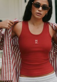 Canotta rossa a coste con ricamo "self.", indossata sotto una camicia a righe con bottoni. La modella porta occhiali da sole scuri, mostrando un look casual.