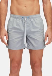 Pantaloni da bagno grigi con vita elastica e cordino, indossati da un uomo in piedi, che mostrano un look casual pronto per la spiaggia.