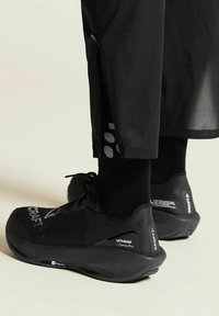 Persoon in zwarte Craft hardloopschoenen, zwarte sokken en zwarte broek met een zijrits op een lichte achtergrond.