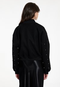 Schwarzer Pullover mit geripptem Kragen und Bündchen, verziert mit dekorativen silbernen Verzierungen an den Ärmeln und einem lässigen Schnitt.