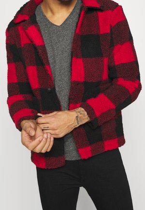 Rot-schwarz karierte Fleecejacke mit kragenförmigem Ausschnitt, einzelner Knopfverschluss und strukturiertem Stoff, getragen über einem grauen V-Ausschnitt-Shirt.