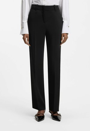 Kvinde iført sorte, højtaljede, skræddersyede bukser, hvid langærmet skjorte med manchetknapper og sorte slingback-sko med spids tå.