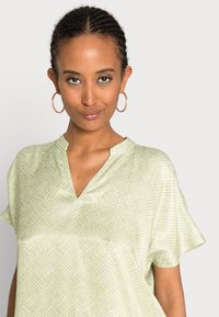 Blusa verde-claro com decote em V, mangas curtas e padrão de quadrados. Tecido suave com um brilho sutil e corte descontraído. Brincos de argola em ouro rosa.
