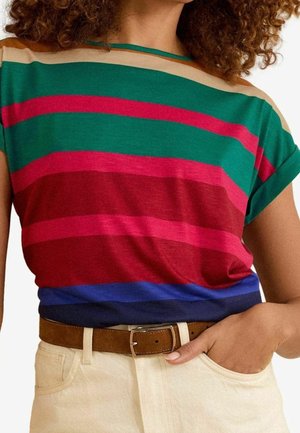 Kobieta w kolorowej, paskowanej koszulce z krótkim rękawem wpuszczonej w beżowe spodnie z brązowym zamszowym paskiem, z ręką włożoną do kieszeni.