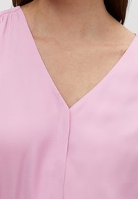 Světle růžová blůza s výstřihem do V, krátkými nabíranými rukávy a hladkým povrchem. Obsahuje středový šev probíhající po přední části.