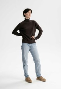 Brauner Rollkragenpullover aus glattem Strickstoff, kombiniert mit hellblauen Jeans und hellbraunen Schuhen, vor einem weißen Hintergrund stehend.