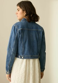 Chaqueta de mezclilla azul con un diseño entallado, detalles de botones y una longitud corta, combinada con un vestido blanco texturizado debajo.