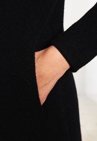 Zwarte trui met lange mouwen, gemaakt van een zachte stof, met een zijzak en een gladde textuur. Close-up van een hand in de zak.