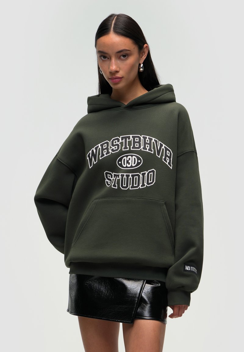 Felpa oversize verde oliva con tasca frontale e testo grafico bianco "WRSTBHVR STUDIO" sul petto. Indossata con una mini gonna nera lucida.