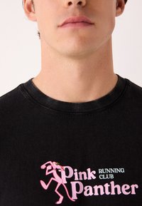 Črna bombažna majica s prazničnim grafikom Pink Panther, z napisom »Pink Panther RUNNING CLUB« v beli in modri barvi. Rebrast izrez ovratnika.