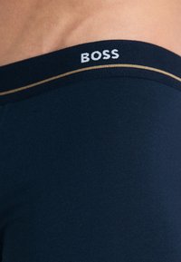 Boxers bleus foncés avec une ceinture élastique marquée "BOSS" en blanc, accentuée par une fine bande dorée. Texture de tissu lisse.