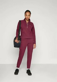 Bordowy strój sportowy z rozpinanym dresowym swetrem i dopasowanymi spodniami, wykonany z elastycznego materiału, połączony z czarną torbą i czarnymi sneakersami.