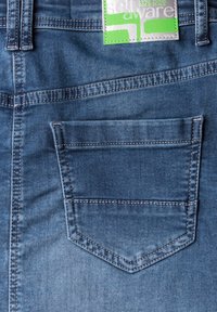 Jeans en denim de couleur bleue délavée, avec une poche arrière dotée d'un rabat arrondi et de coutures contrastantes. L'étiquette affiche les détails de la marque en vert.