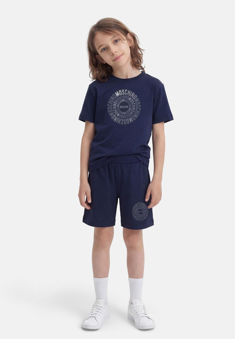 Otrok z ramenoma dolgimi lasmi, oblečen v temno modro majico in kratke hlače Moschino ter bele nogavice in superge, stoji na belem ozadju.
