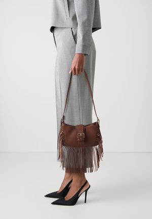 Persona in completo a quadri monocromo che tiene una borsa a frange in pelle marrone, indossando décolleté slingback nere con punta appuntita, in piedi su sfondo bianco.