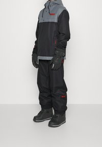 Mustan ja harmaan värinen hiihtotakki, jossa on huppu, joustavat hihansuut ja etutasku. Yhdistetty löysiin mustiin housuihin ja mustiin vedenpitäviin saappaisiin.