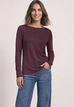 Femme aux cheveux bruns mi-longs portant un haut à manches longues violet foncé et un jean bleu, debout avec une main dans la poche et souriante.