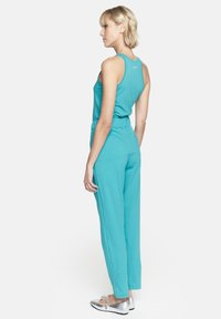 Turkusowy kombinezon bez rękawów, z elastycznym pasem i szerokimi nogawkami. Materiał wydaje się miękki, ma swobodny krój i nie ma widocznych wzorów.