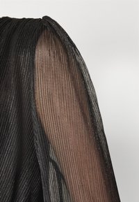 Tissu noir plissé transparent avec un léger scintillement, drapé sur une épaule et un bras sur un fond clair uni.