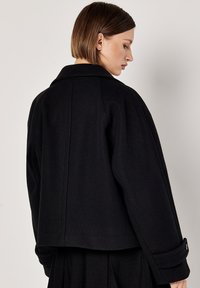 Giacca in lana nera con colletto a scollo a V, vestibilità oversize e dettagli sui polsini con bottoni. Texture liscia e design pulito e minimal.