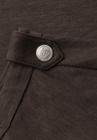 Tissu texturé marron foncé avec un bord plié maintenu par un bouton argenté présentant un emblème de couronne et de bouclier.