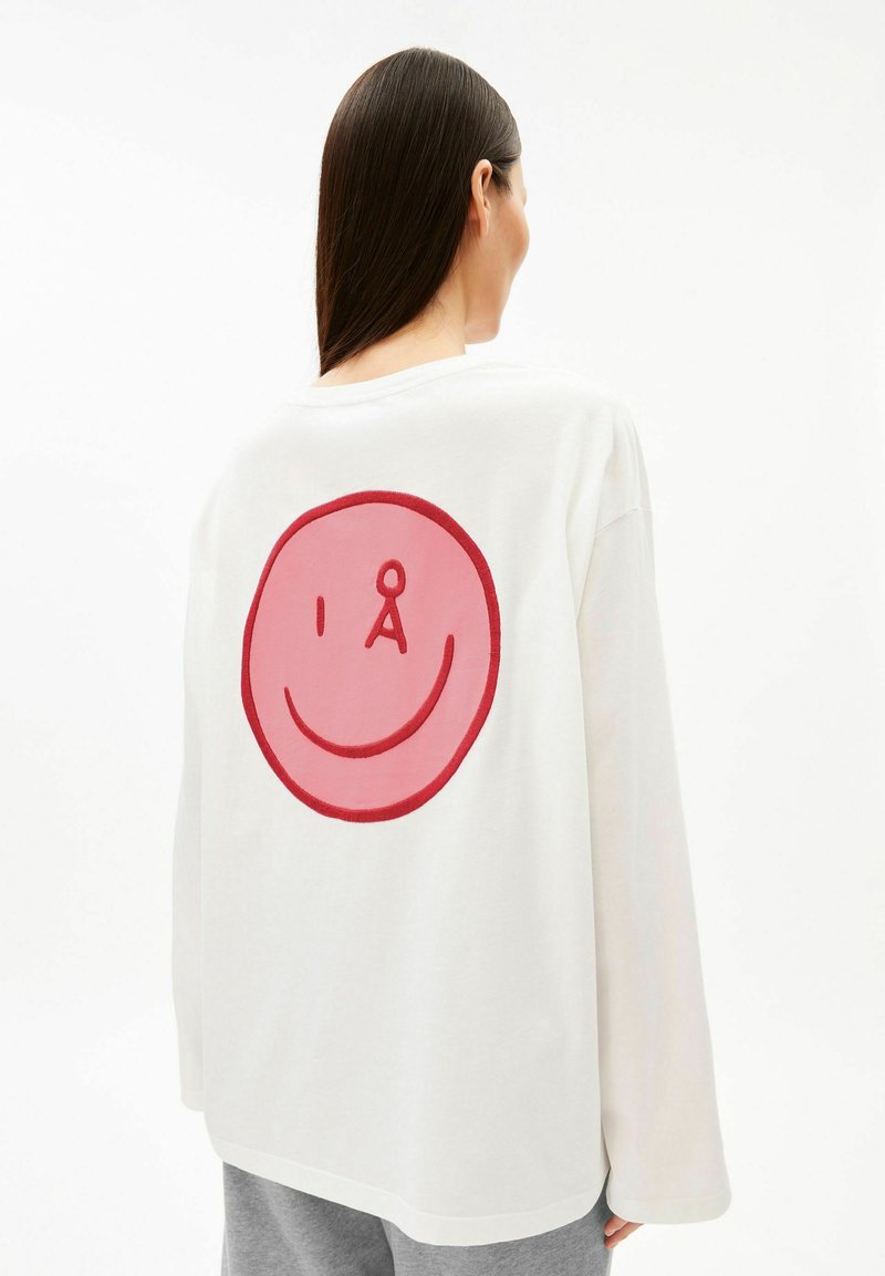 Person mit langen dunklen Haaren, die von hinten zu sehen ist, trägt ein weißes Langarmshirt mit großem roten und rosa Smiley-Gesicht auf dem Rücken.