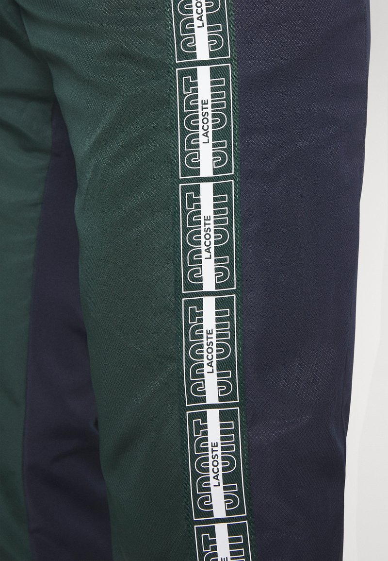 Pantalons verts et bleu marine avec une surface texturée, présentant de grands logos blancs "SPORT" sur le côté et un design contrasté.