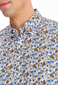 Biała koszula na guziki z kwiatowym wzorem, z niebieskimi i pomarańczowymi kwiatami, liściastymi akcentami oraz fakturowaną tkaniną.