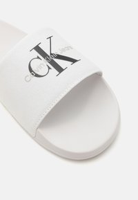 Hvit glidesandal med en stoffstropp som viser "Calvin Klein Jeans" i svart. Glatt gummisåle og strukturert stroppoverflate.