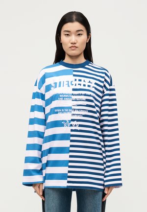 Γυναίκα που φοράει μπλούζα με μακριά μανίκια, άνισες οριζόντιες ρίγες σε μπλε και άσπρο χρώμα και τυπωμένο κείμενο, στέκεται μπροστά από μια απλή φόντο.