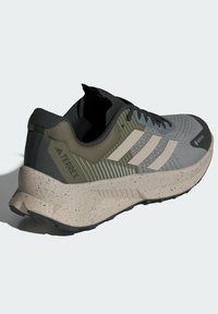 Grå och grön träningssko med en texturerad ovandel, tre vita ränder och en prickig beige sula. Har förstärkt häl och snörningsdesign.