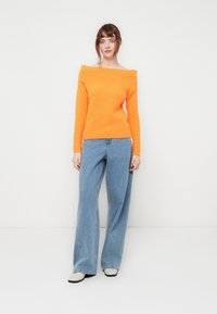 Živahna oranžna pletena majica z off-the-shoulder krojem je v kombinaciji z svetlo modrimi širokimi hlačami. Majica ima rebrast teksturo, hlače pa so sproščenega kroja.