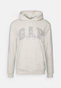 Sweat à capuche beige clair avec de grandes lettres grises "GAP" sur la poitrine, poche kangourou à l'avant et capuche avec cordon de serrage.