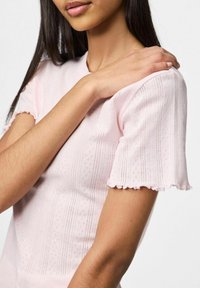 Jasnoróżowa bluzka z krótkim rękawem o teksturowanym wzorze i falbaniastych krawędziach. Materiał wydaje się miękki z subtelnym paskowanym wzorem i małymi perforacjami.
